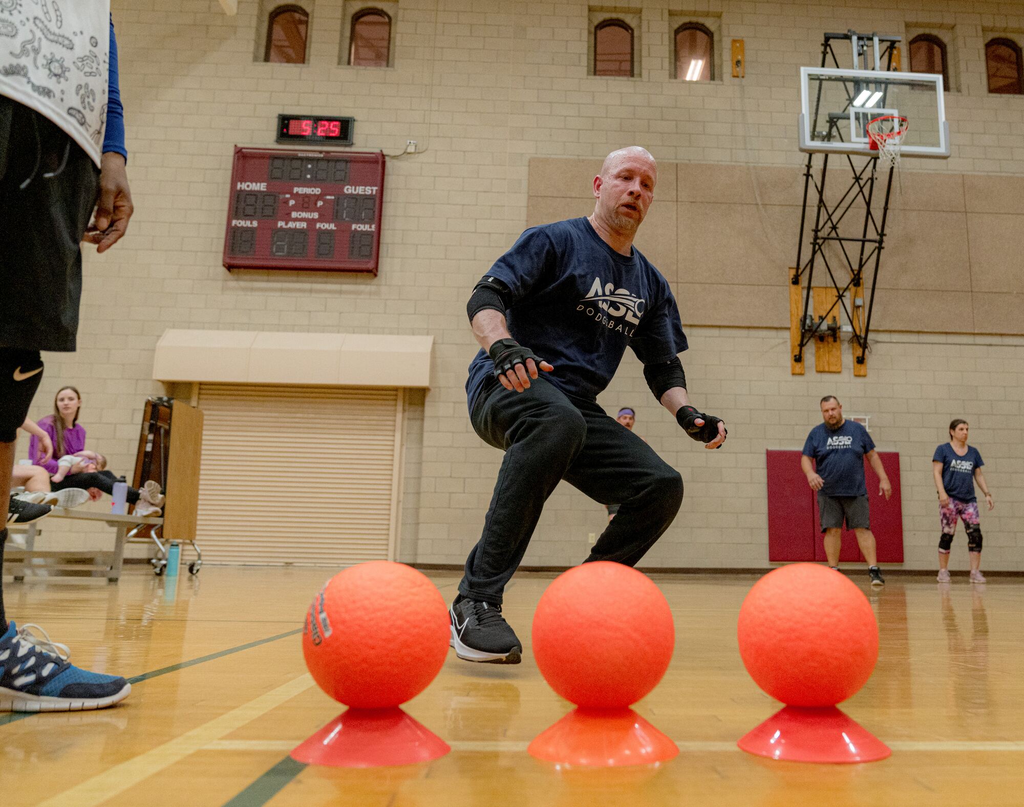 A player crouched at the center line of the gym, three orange dodgeballs on cones in front of him, waiting for the opening rush